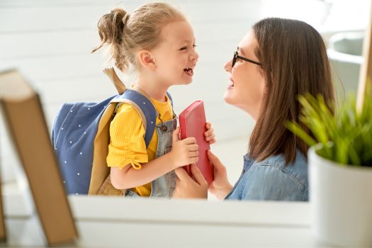 Mother smiling and bending down to eye level with her young happy daughter handing her a book to add to her backpack on her back