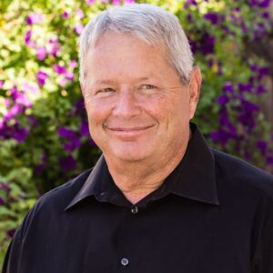 Warren Kohn in a black shirt smiling in front of purple flowers